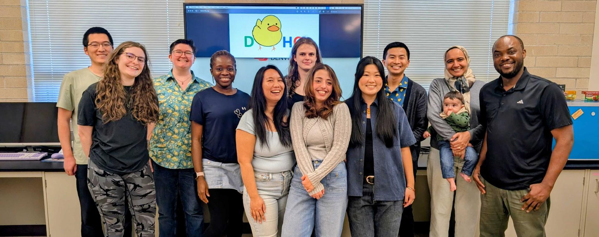 A picture of the DUHCI group, with several students and professors smiling at the camera in front of a sign with the DUHCI duck-themed logo.