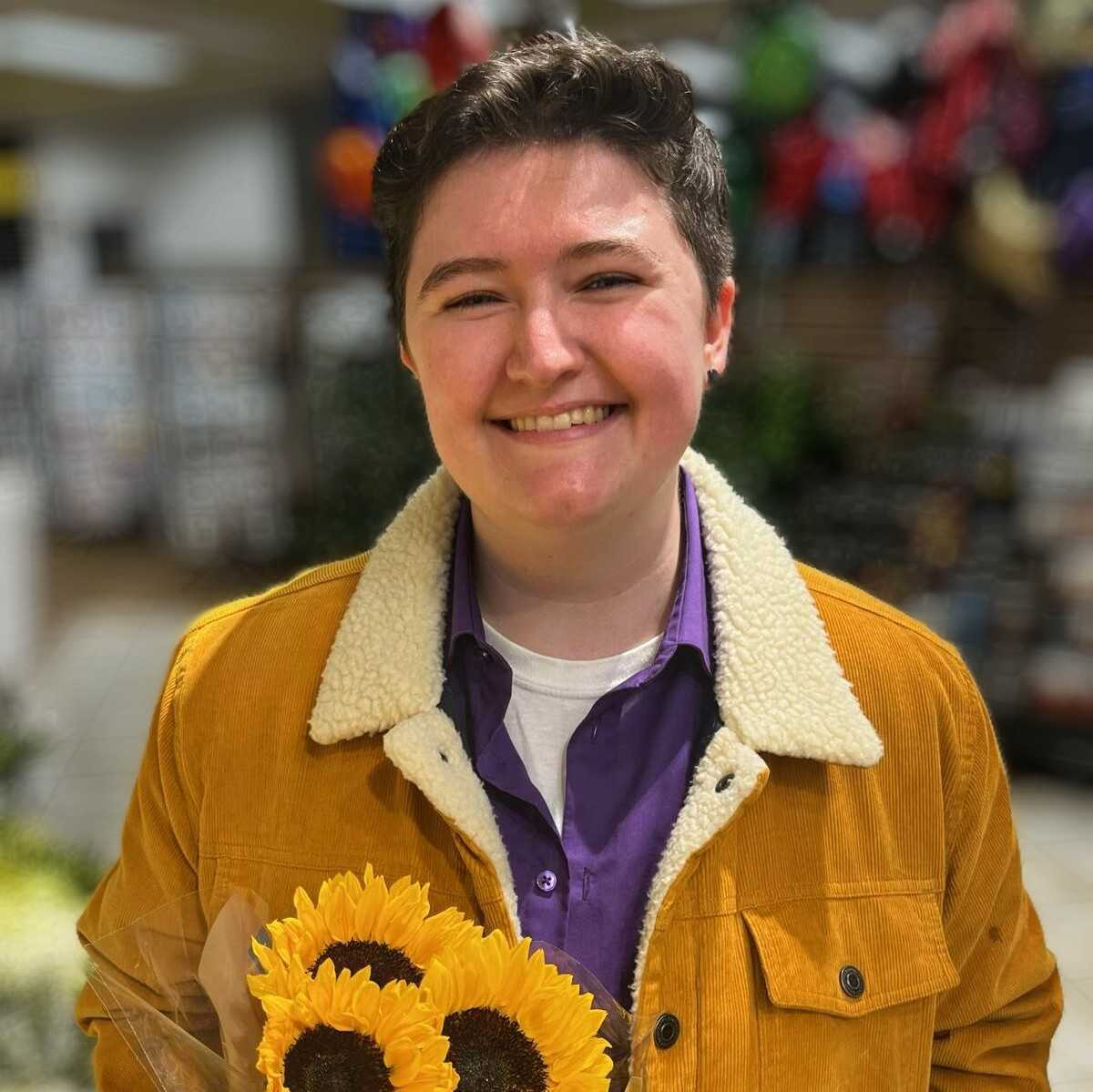 A late 20s person with short dark hair, a bright yellow jacket, and a purple button down shirt smiling while holding a bouquet of sunflowers.